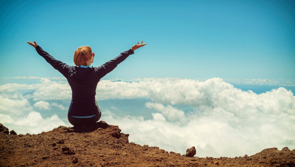 person sitting on cliff raising up both hands