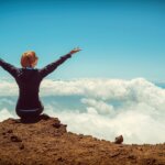 person sitting on cliff raising up both hands